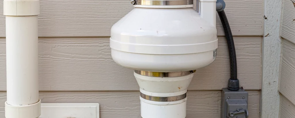 Exterior residential radon mitigation system fan and PVC venting pipe mounted on house siding.